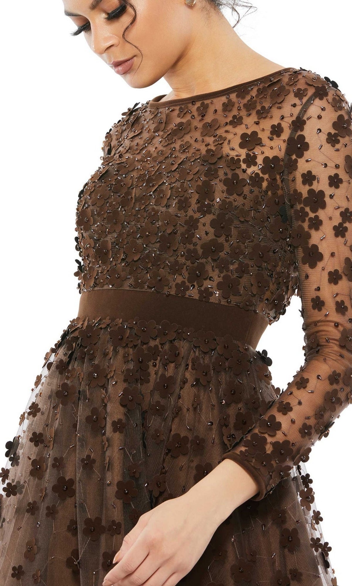 A woman models the Mac Duggal 67007 knee-length wedding-guest dress, featuring sheer brown fabric, floral embellishments with 3D appliqués, long sleeves, and a fitted waistband. She looks down with her hand near her waist.
